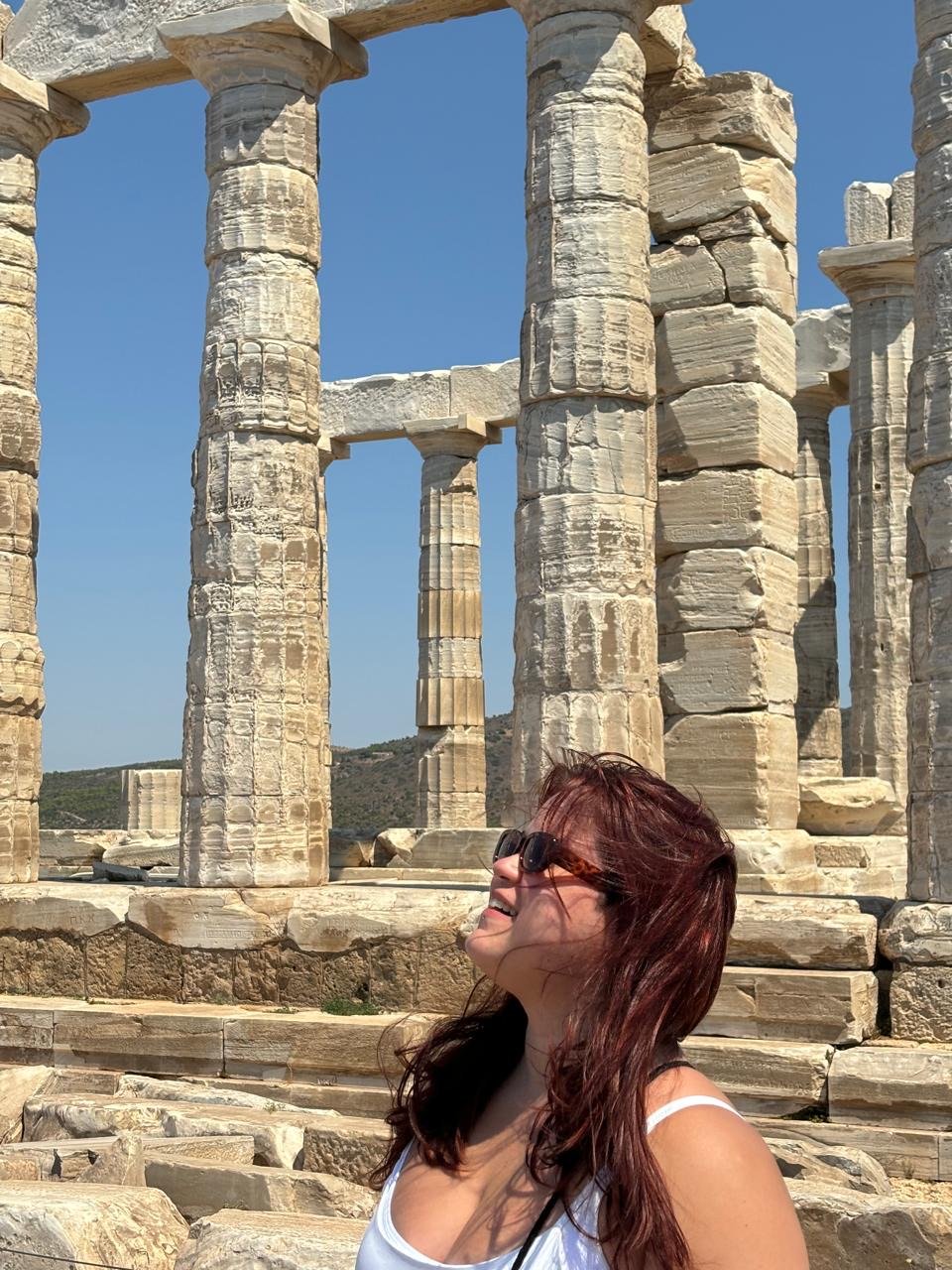 Yrini sorrindo e olhando para cima diante das colunas do Templo de Poseidon, em Sunion, na Grécia